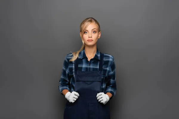 Femme de travail en gants et salopettes regardant la caméra isolée sur gris — Photo de stock