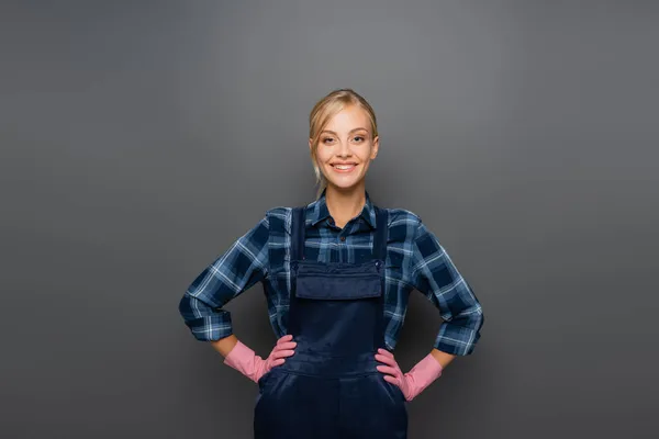 Happy workwoman in rubber gloves and overalls looking at camera on grey — Stock Photo