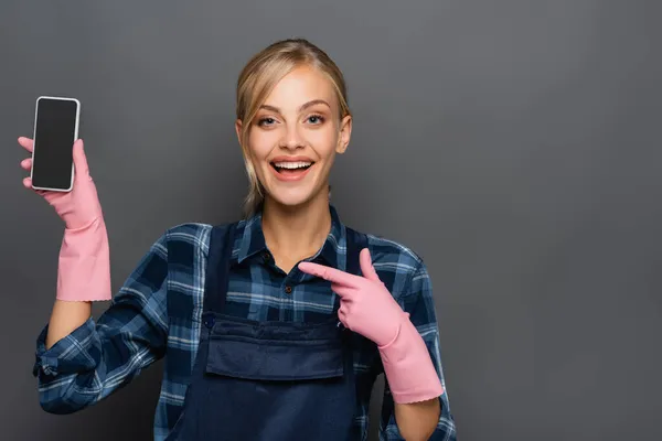 Alegre fontanero rubio en guantes de goma apuntando al teléfono inteligente con pantalla en blanco aislado en gris - foto de stock