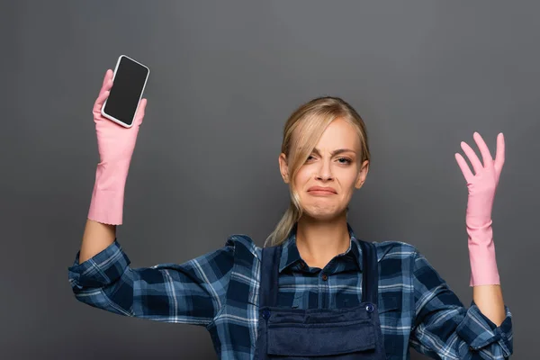 Fontanero trastornado en mono y guantes de goma que sostiene el teléfono inteligente con pantalla en blanco aislado en gris - foto de stock