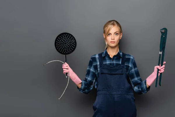 Pensive plumber in rubber gloves holding pipe wrench and shower head on grey — Stock Photo