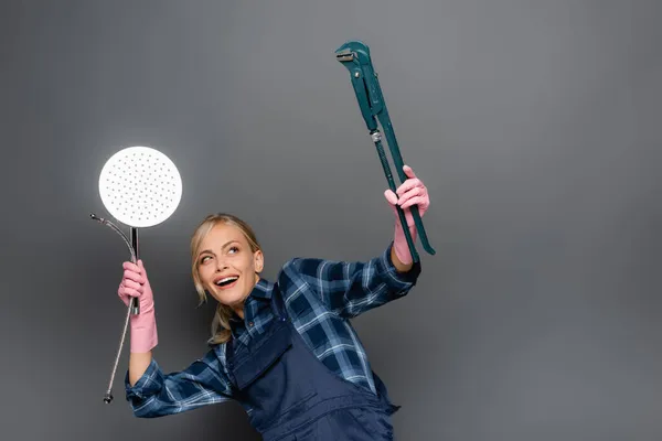 Positive plumber in rubber gloves and overalls holding shower head and looking at pipe wrench isolated on grey — Stock Photo