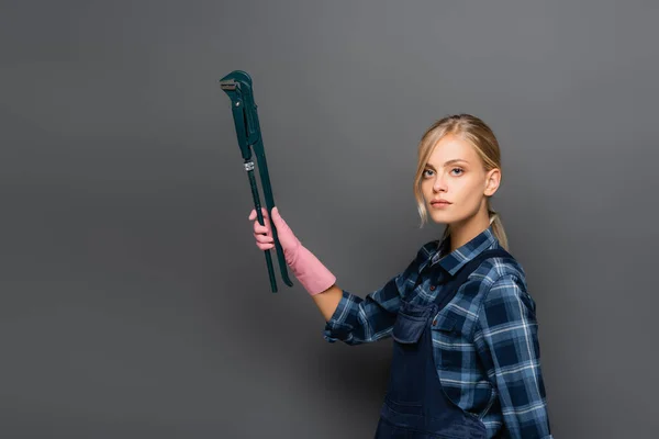 Blonde plumber holding pipe wrench and looking at camera on grey background — Stock Photo