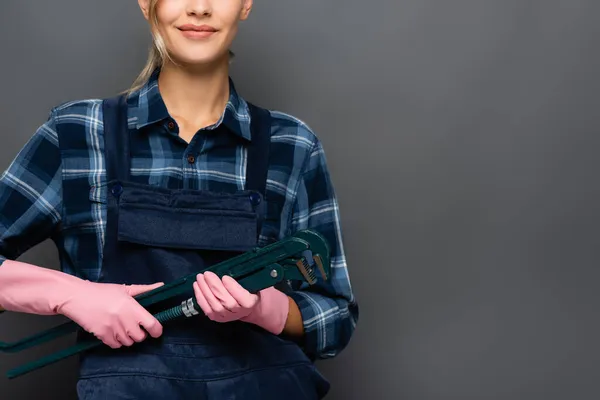 Vista ritagliata di idraulico sorridente in tuta e guanti di gomma tenendo chiave del tubo isolato su grigio — Foto stock