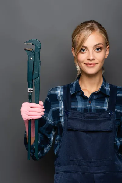 Young blonde plumber in plaid shirt and overalls holding pipe wrench on grey background — Stock Photo