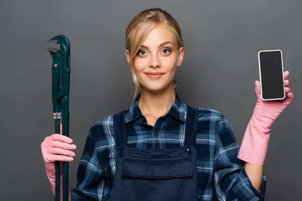 Blonder Klempner in Overalls hält Handy mit leerem Bildschirm und Rohrschlüssel isoliert auf grau — Stockfoto