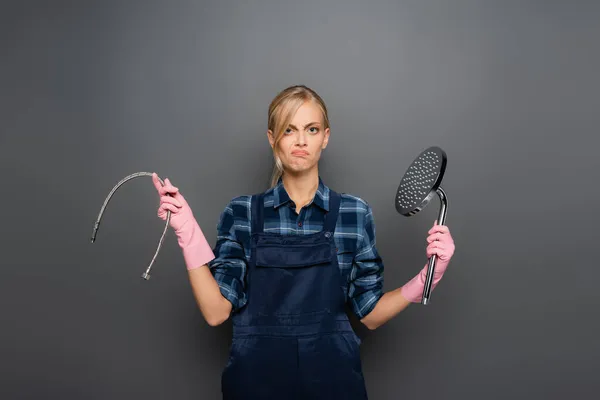 Displeased plumber in rubber gloves holding pipe and shower head on grey background — Stock Photo