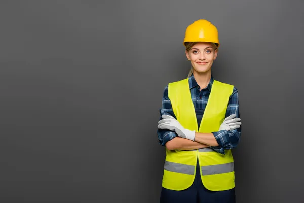 Construtor sorridente em hardhat de pé com braços cruzados isolados em cinza — Fotografia de Stock