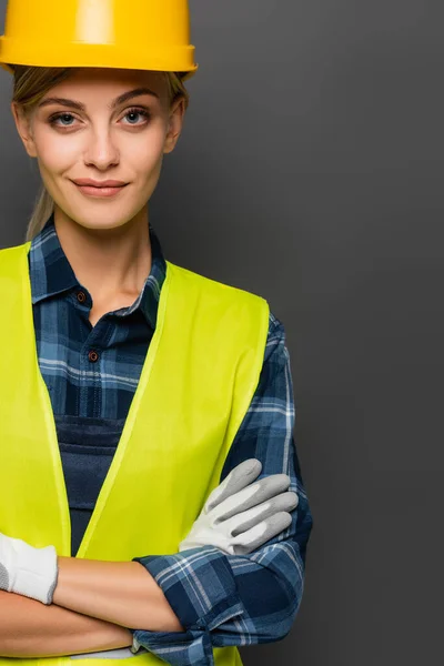 Smiling builder in helmet and safety vest crossing arms isolated on grey — Stock Photo