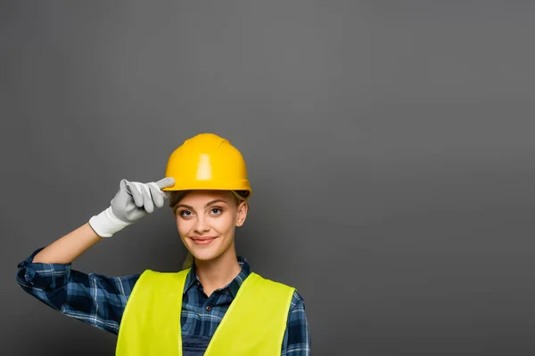 Constructor positivo en casco y chaleco de seguridad mirando a la cámara aislada en gris - foto de stock