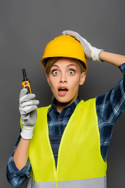 Constructor asustado en hardhat y chaleco de seguridad sosteniendo walkie talkie y mirando a la cámara aislada en gris - foto de stock