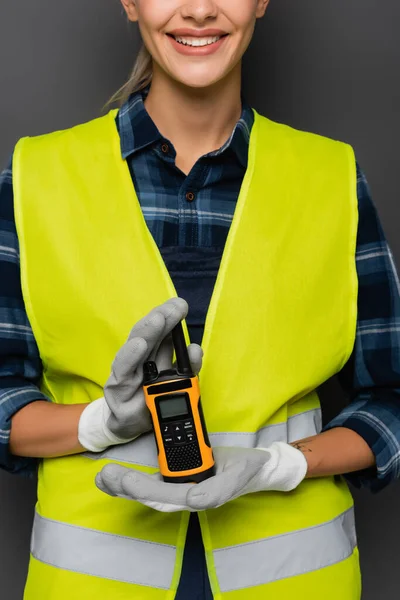 Vista cortada do construtor positivo em colete de segurança e luvas segurando walkie talkie isolado em cinza — Fotografia de Stock