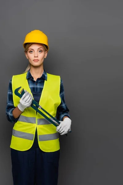 Blonde Bauarbeiterin in festem Hut mit Rohrschlüssel und Blick auf Kamera isoliert auf grau — Stockfoto