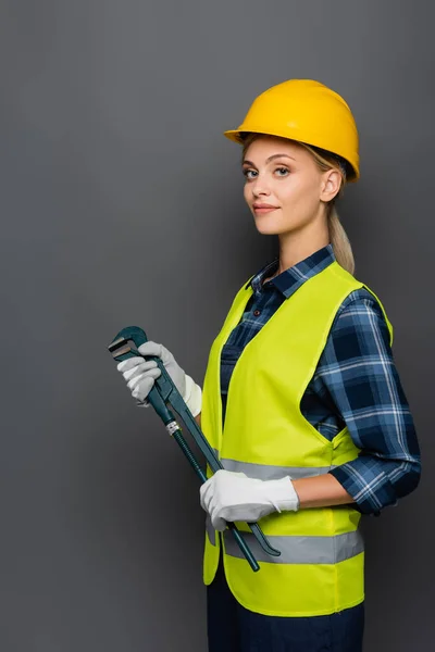 Jeune constructeur en casque et gilet de sécurité tenant la clé de tuyau isolé sur gris — Photo de stock