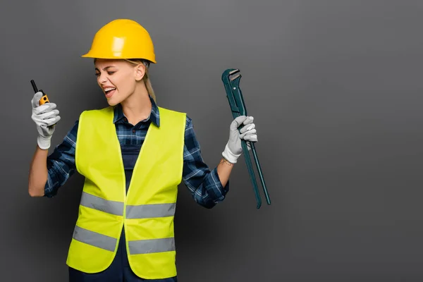 Construtor irritado em capacete gritando em walkie talkie e segurando chave de tubo isolado em cinza — Fotografia de Stock