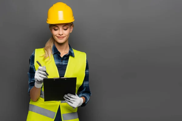 Constructor sonriente en hardhat y chaleco de seguridad que sostiene la pluma y el portapapeles aislados en gris - foto de stock