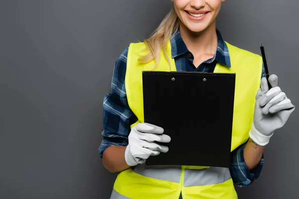 Cropped view of cheerful blonde builder in gloves and safety vest holding pen and clipboard isolated on grey — Stock Photo