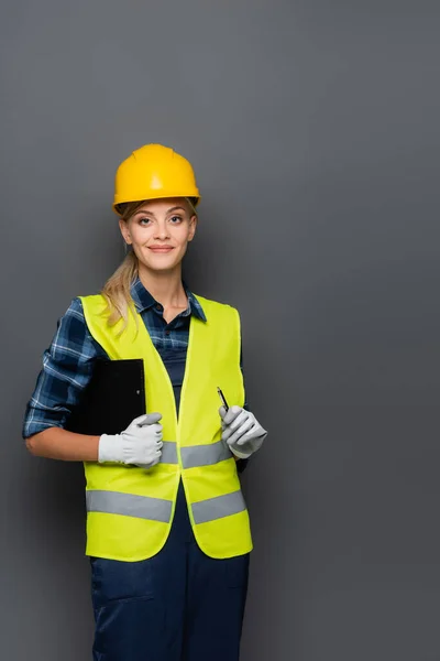 Allegro costruttore in hardhat guardando la fotocamera mentre tiene appunti e penna isolati sul grigio — Foto stock