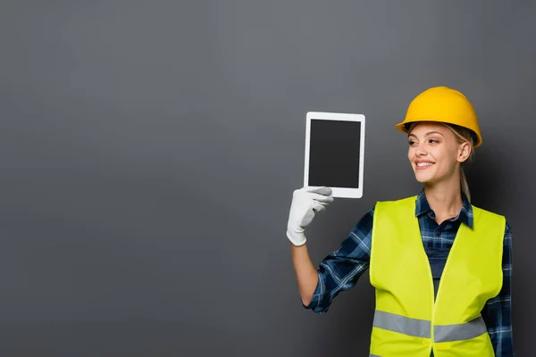 Construtor loira sorridente em chapéu duro segurando tablet digital com tela em branco isolado em cinza — Fotografia de Stock