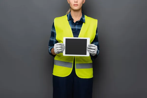 Cropped view of builder in safety vest and gloves holding digital tablet with blank screen isolated on grey — Stock Photo