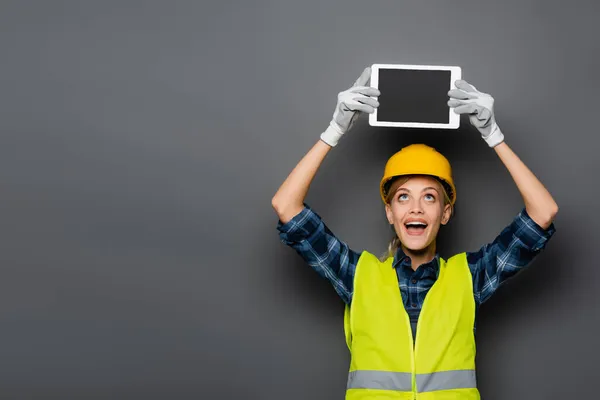 Emocionado constructor de guantes y casco que sostiene la tableta digital con pantalla en blanco aislado en gris - foto de stock