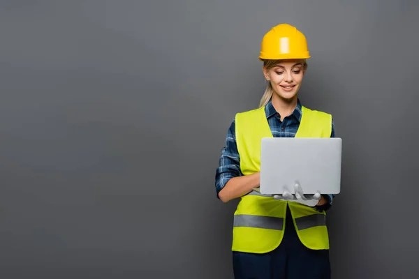 Positive builder in hardhat and protective vest using laptop isolated on grey — Stock Photo