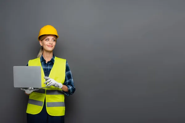 Fröhlicher Bauarbeiter in Handschuhen und Helm, Laptop isoliert auf grau — Stockfoto