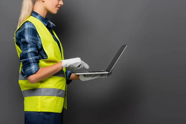 Cropped view of blonde builder in safety vest and gloves pointing at laptop isolated on grey — Stock Photo