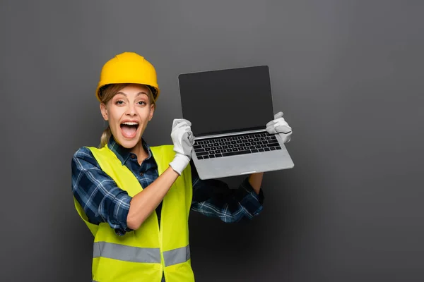 Emocionado constructor en chaleco de seguridad que sostiene el ordenador portátil con pantalla en blanco aislado en gris - foto de stock