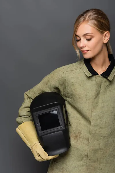 Bella bionda saldatore in uniforme guardando casco isolato su grigio — Foto stock