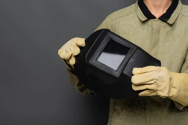 Vista recortada del soldador en uniforme y guantes que sostienen la máscara sobre fondo gris - foto de stock