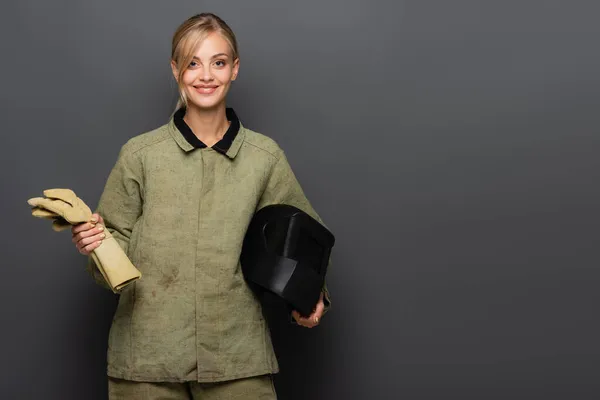 Cheerful blonde welder holding gloves and protective helmet on grey background — Stock Photo