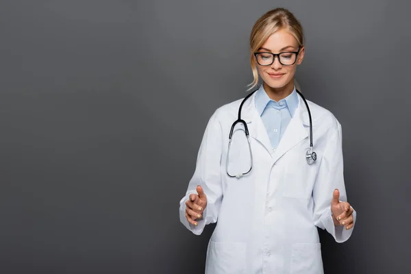 Smiling blonde doctor gesturing and looking down isolated on grey — Stock Photo