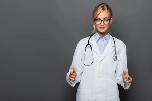 Sorridente medico bionda gesticolando e guardando giù isolato sul grigio — Foto stock