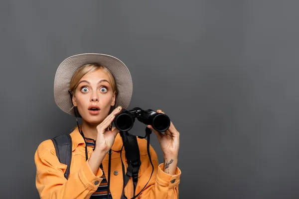 Shocked tourist with backpack holding binoculars and looking at camera isolated on grey — Stock Photo