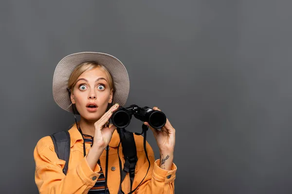 Turista sorprendido con mochila sosteniendo binoculares y mirando a la cámara aislada en gris - foto de stock