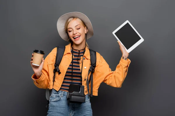 Happy tourist with backpack and vintage camera holding digital tablet and paper cup on grey background — Stock Photo