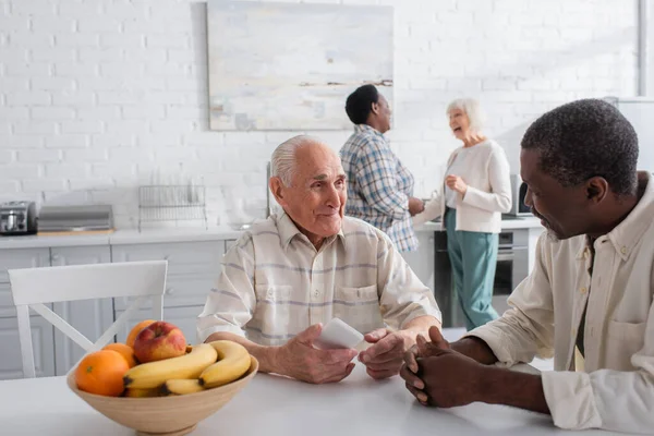 Sorrindo homens inter-raciais seniores com smartphone falando perto de frutas em casa de repouso — Fotografia de Stock