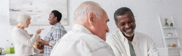 Smiling elderly interracial men talking near blurred women in nursing home, banner — Stock Photo