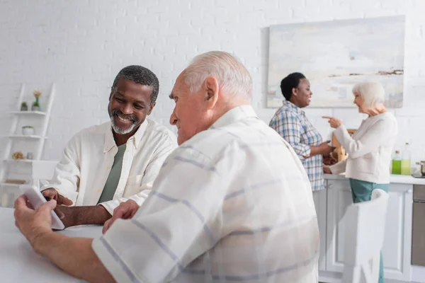 Alegre hombres mayores utilizando el teléfono inteligente cerca borrosa amigos multiétnicos en el hogar de ancianos - foto de stock