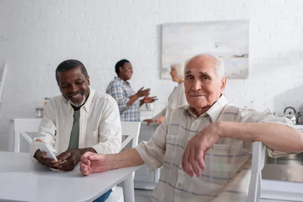 Пожилой мужчина смотрит в камеру рядом с улыбающимся африканским американским другом со смартфоном в доме престарелых — стоковое фото