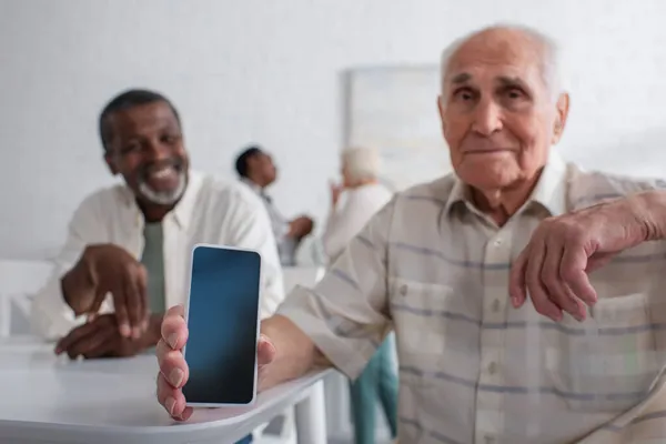 Smartphone en la mano de un hombre mayor borroso cerca de un amigo afroamericano en un hogar de ancianos — Stock Photo