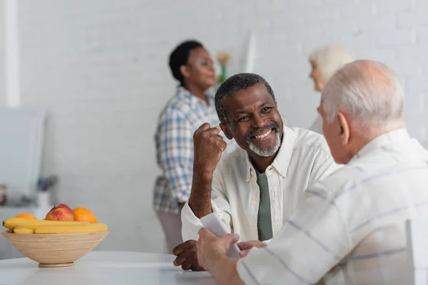Sonriente hombre afroamericano mostrando sí gesto cerca de amigo mayor con teléfono inteligente y mujeres borrosas en el hogar de ancianos - foto de stock