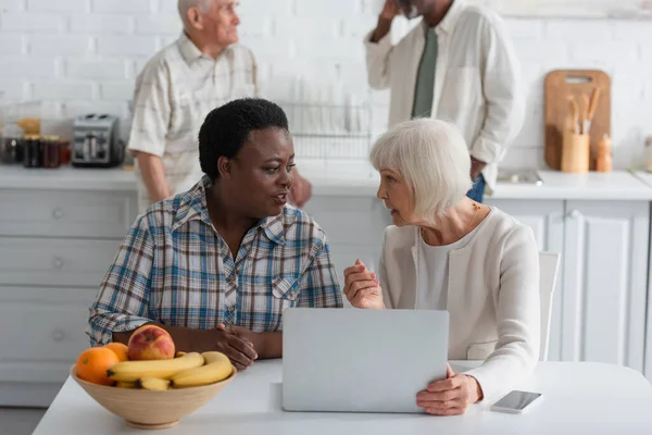 Senior interracial mulheres falando perto de dispositivos e frutas em casa de repouso — Fotografia de Stock