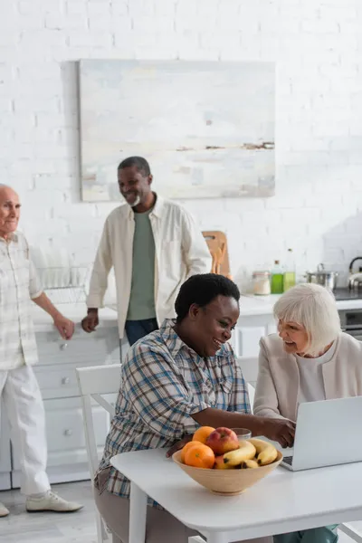 Souriantes femmes multiethniques utilisant un ordinateur portable près des fruits dans une maison de retraite — Photo de stock