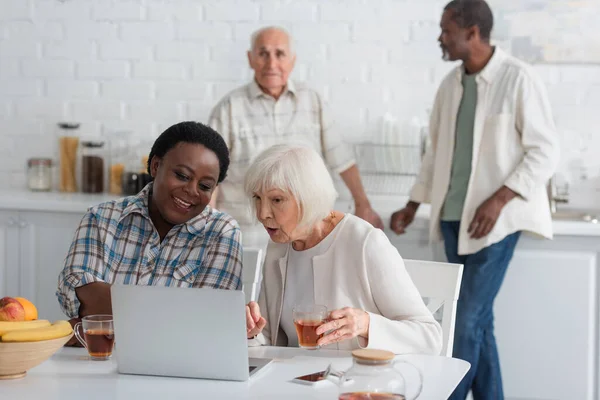 Mayores mujeres multiétnicas con té usando portátil cerca de frutas en el hogar de ancianos - foto de stock