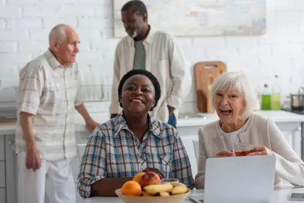 Cheerful multiethnic women with tea looking at camera near devices and fruits in nursing home — Stock Photo