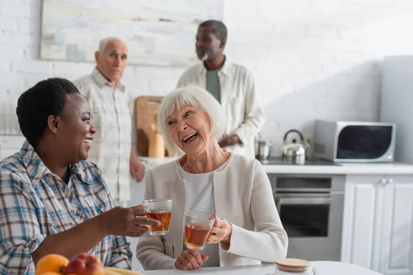 Feliz sénior interracial mujeres celebración de té cerca de frutas en el hogar de ancianos — Stock Photo