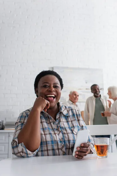 Happy african american woman holding smartphone near tea in nursing home — Stock Photo