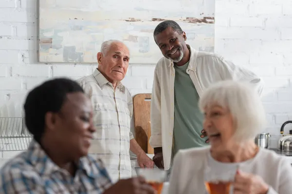 Hombres multiétnicos mayores mirando a la cámara cerca de mujeres sosteniendo té en un asilo de ancianos - foto de stock